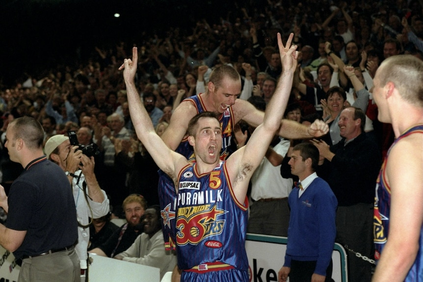 A basketball player with his hands up in a V pose while another man walks on seats among a cheering crowd