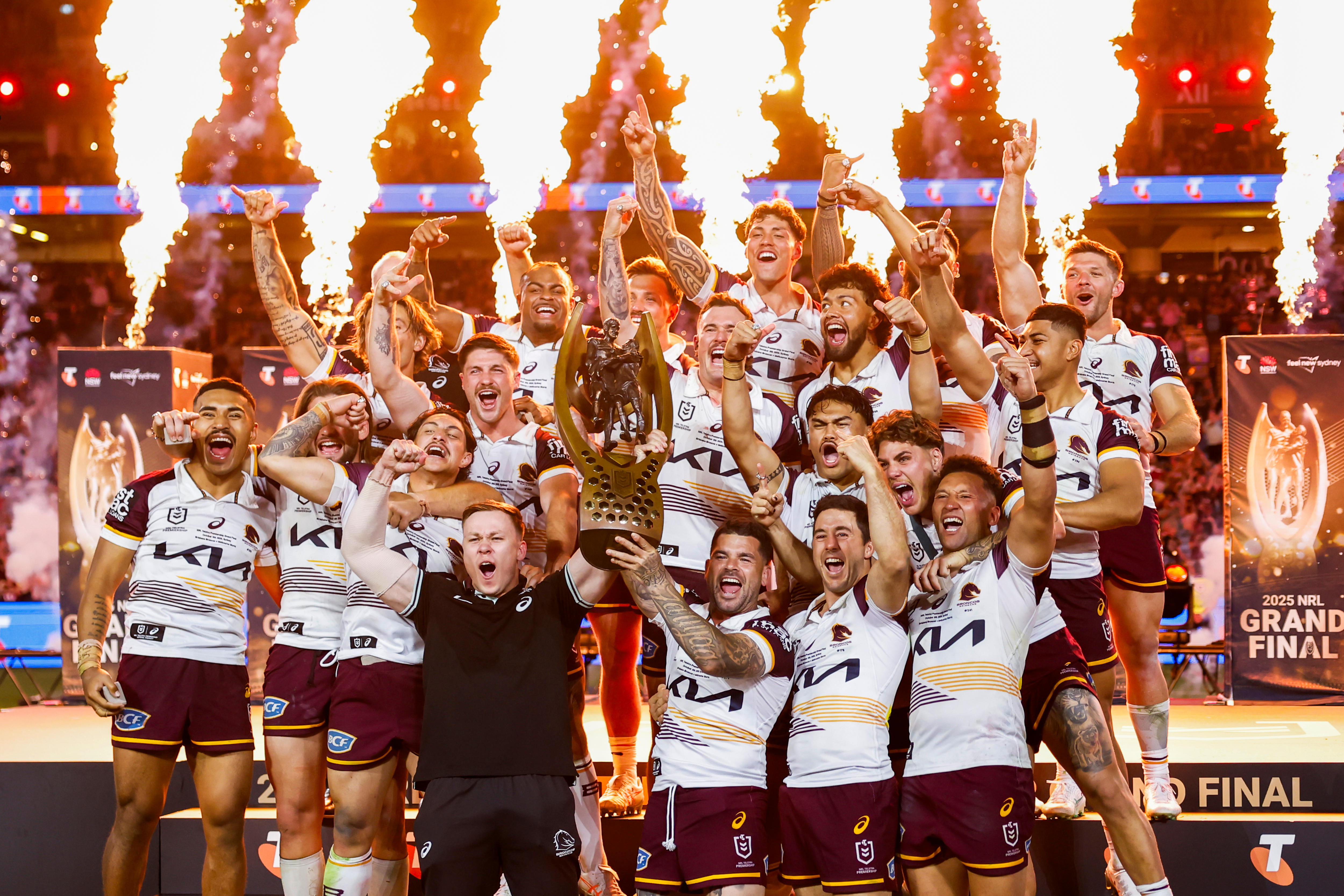 Jogador do Brisbane Broncos torcendo com os braços levantados e levantando o troféu da NRL em frente a fogos de artifício em chamas