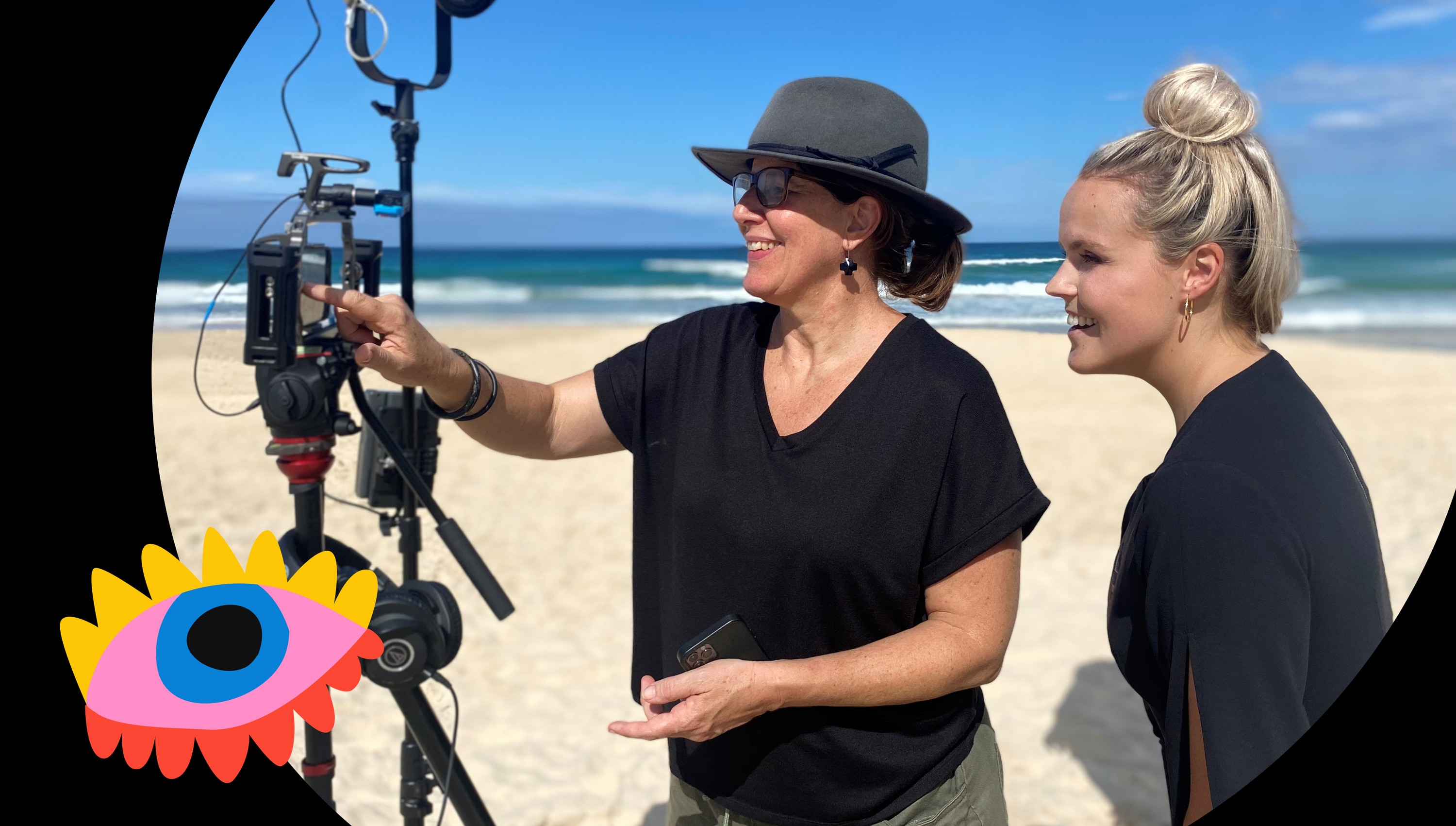  Middle aged woman showing younger woman how to use a mobile phone journalism kit. They are located on a beach.