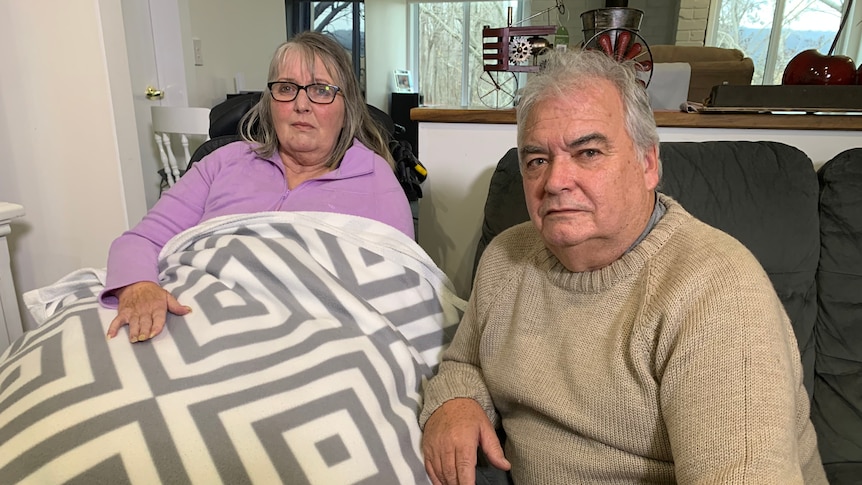 A woman lying in a bed, with her male carer seated next to her.