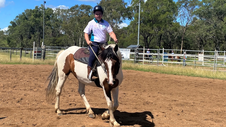 A young woman leads a pinto horse around a traffic cone in a dusty ring under a bright blue sky