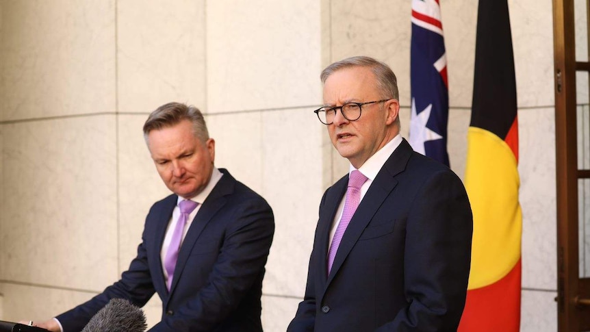 Two men in suits stand in front of flags as they address media.