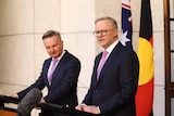 Two men in suits stand in front of flags as they address media.