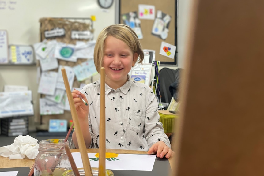 A smiling girl with blond hair and a button down shirt is holding a paintbrush in the air