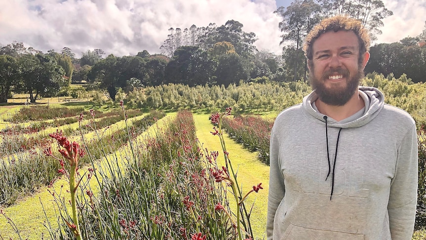 A man with a beard smiles at the camera with rows of kangaroo paws behind him.