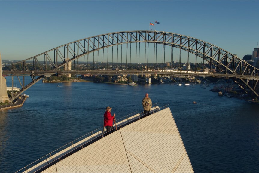 Two men standing on top of the Sydney Opera House with the Harbour Bridge in the background with Aboriginal and Australian flags