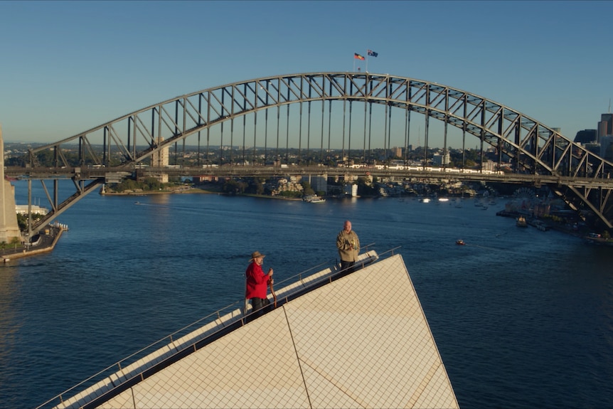 Two men standing on top of the Sydney Opera House with the Harbour Bridge in the background with Aboriginal and Australian flags