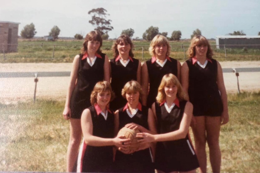 Old photo of seven woman wearing black, red and white netball outfits standing outside.