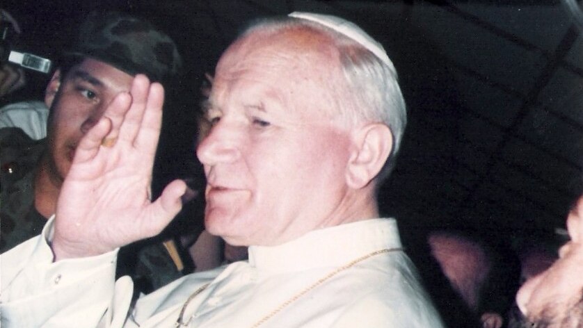 A vintage printed photo shows Pope John Paul II in white vestments waving in front of a media throng.