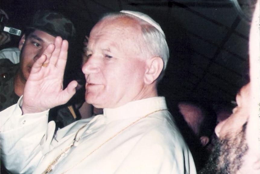 A vintage printed photo shows Pope John Paul II in white vestments waving in front of a media throng.