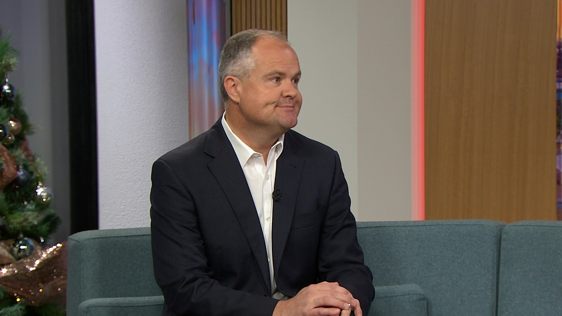 A man in a black suit and white tie seated on a couch in a television studio.