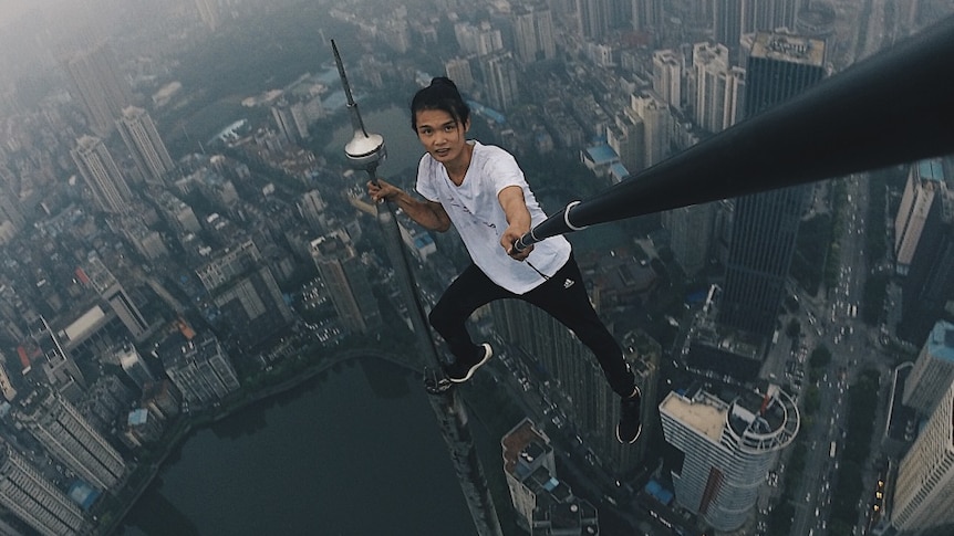 A man holds on to an antenna atop a skyscraper and takes a selfie using a selfie stick