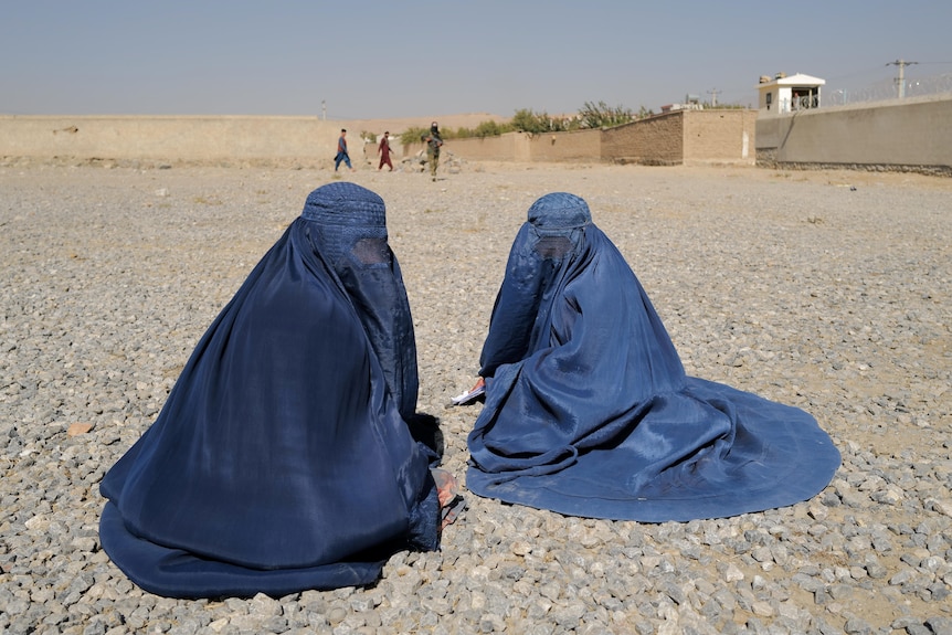Two women in full-face coverings sit on the ground in an arid landscape