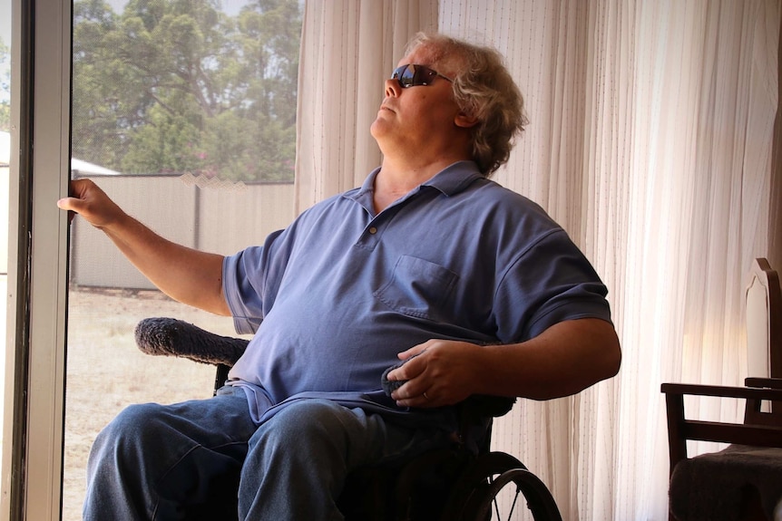 A man in a wheelchair sits in front of a sliding door looking out to a yard