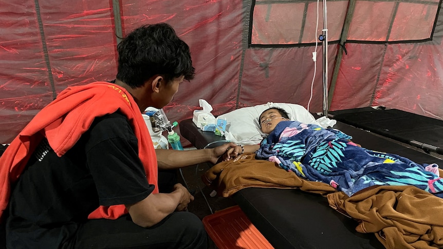 A little boy lays in a makeshift hospital bed inside a tent, with a grown man sitting next to him holding his hand