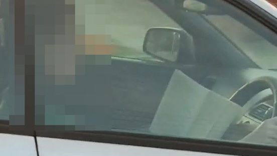 A person, whose face has been pixellated, with a book in the driver's seat of a car.