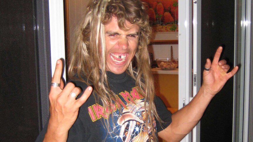 Adam Coleman stands with an Iron Maiden t-shirt and both hands in a rock horns gesture.