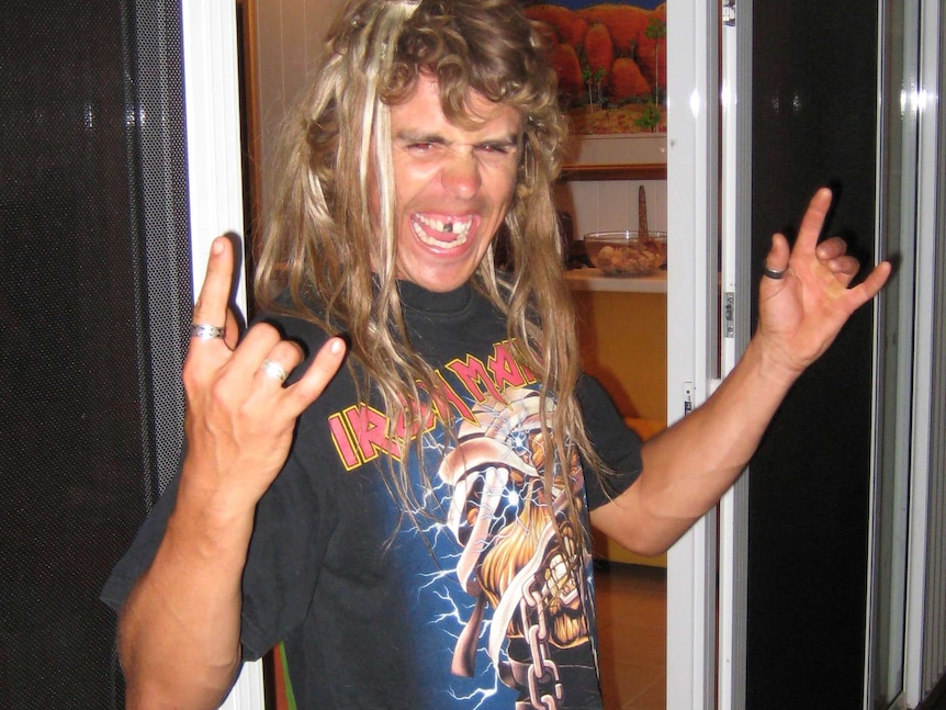 Adam Coleman stands with an Iron Maiden t-shirt and both hands in a rock horns gesture.
