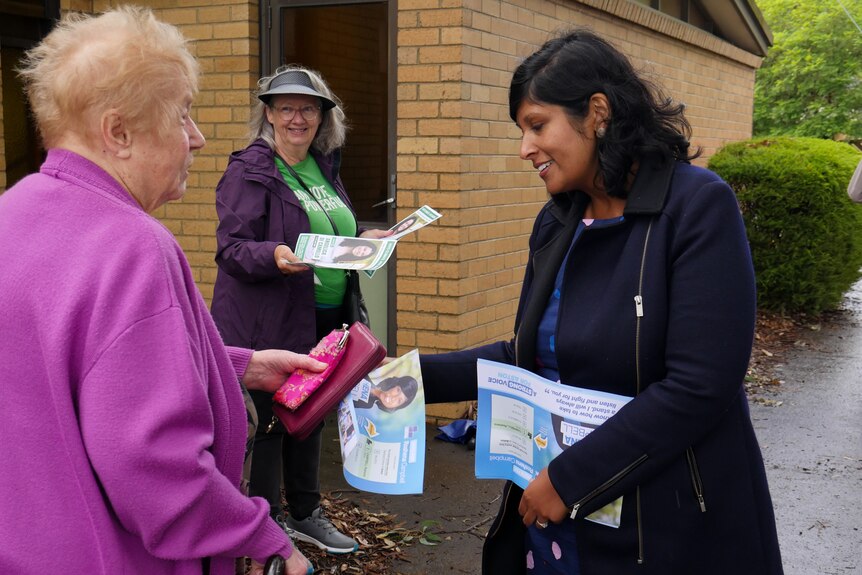 A woman in a blue dress and dark blue jacket hands a flyer to a woman in a purple cardigan 