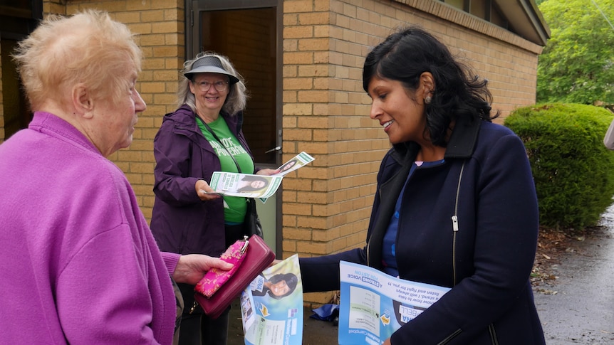 A woman in a blue dress and dark blue jacket hands a flyer to a woman in a purple cardigan 