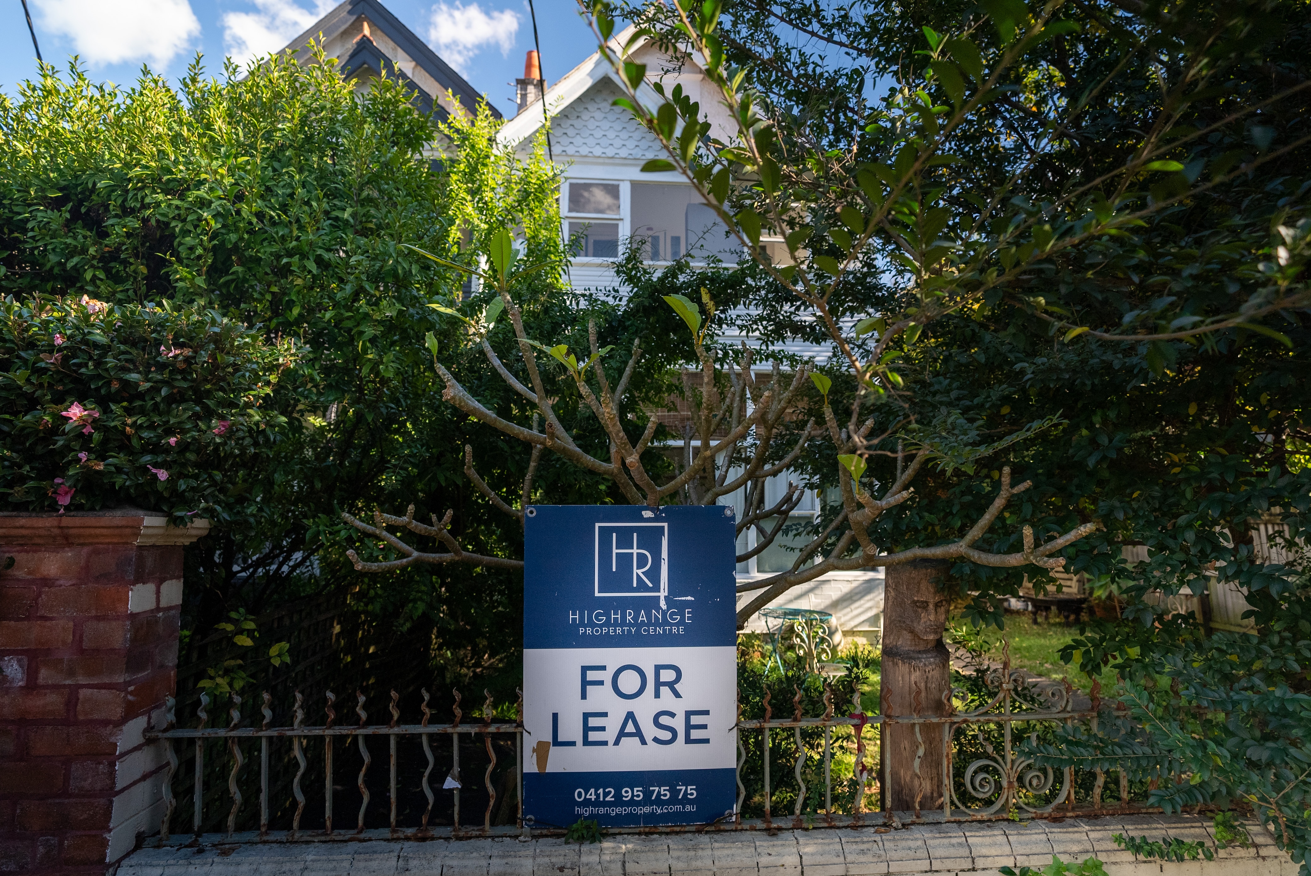 A blue and white for lease sign is mounted onto a metal fence in front of a house surrounded by green foliage.