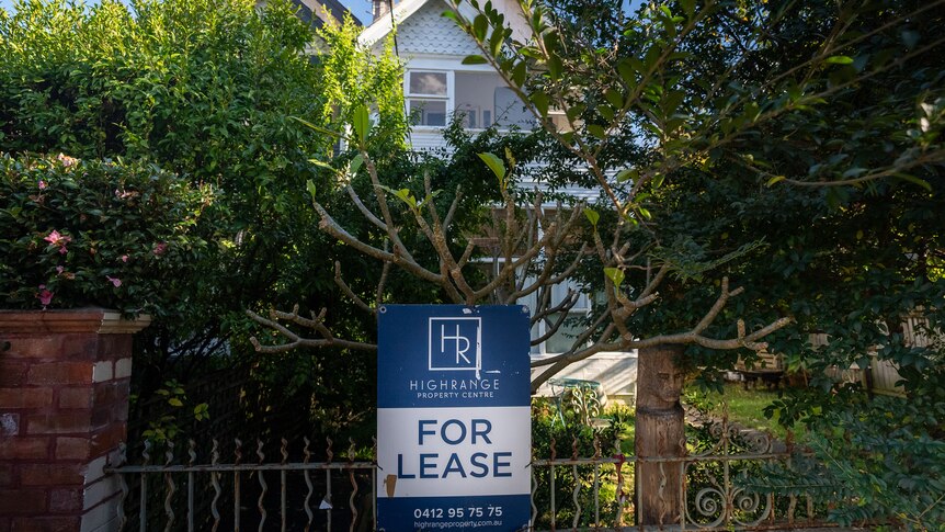 A blue and white for lease sign is mounted onto a metal fence in front of a house surrounded by green foliage.