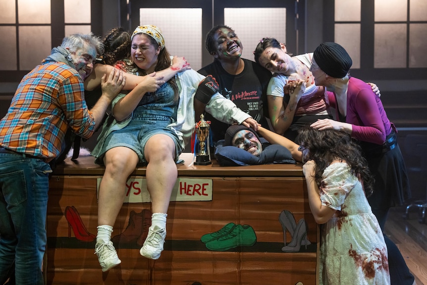 Eight amateur musical theatre performers - some dressed as zombies - embrace onstage.