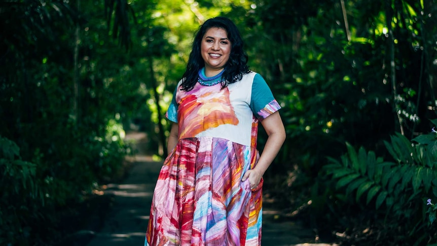 A woman smiles in the middle of a well-lit path, she is wearing a vibrant, patterned dress