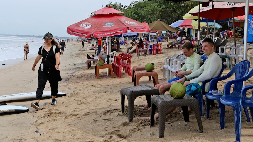 Tourists sit on a beach with a watermellon while a woman walks past.