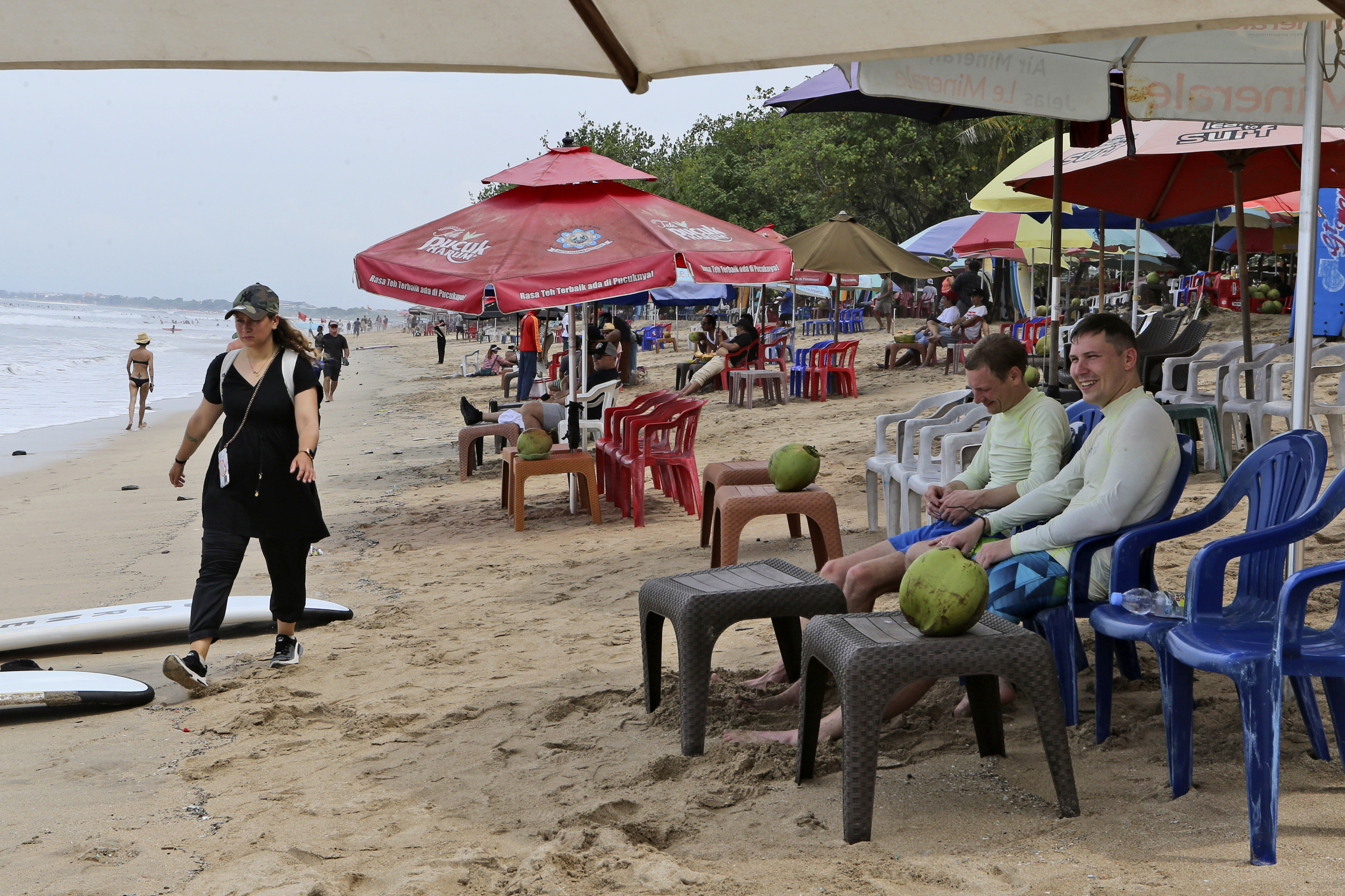 游客拿着西瓜坐在海滩上，而一名妇女从身边走过。