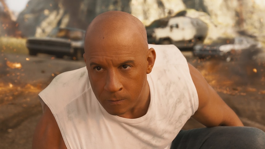 Vin Diesel crouched in front of three cars, with a determined expression, in Fast and Furious 9