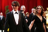 A man in a black tuxedo wearing glasses holds hands with a woman, who is wearing a black dress and looking at him.