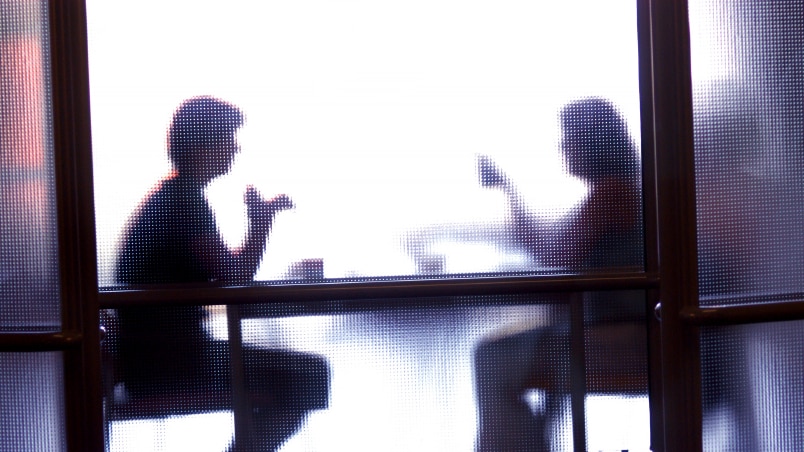 Two people obscured by opaque glass, sit at table.