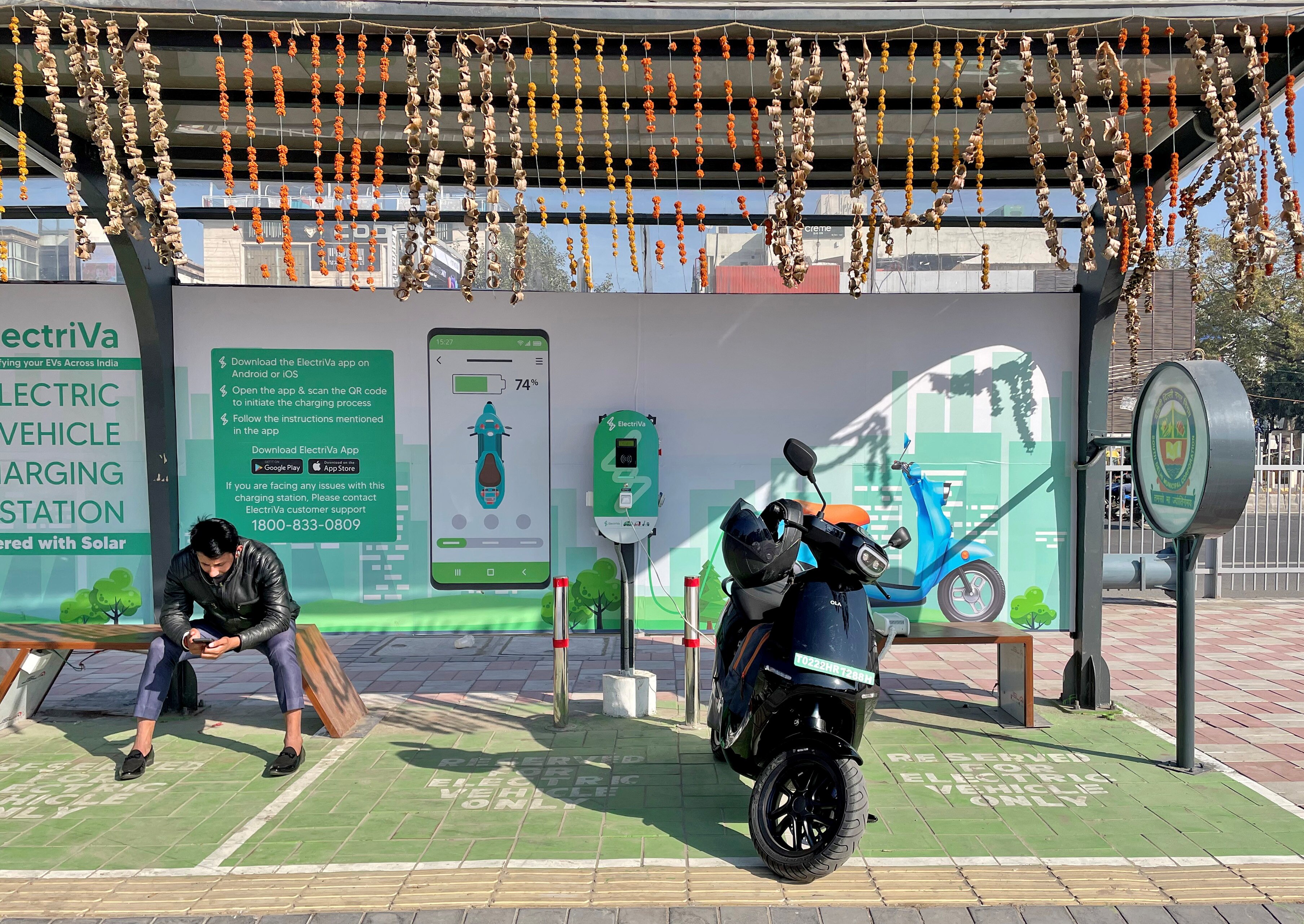 A man checks his mobile phone as he waits while recharging his Ola electric scooter at an electric vehicle charging station