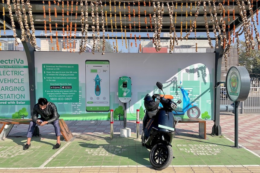 A man checks his mobile phone as he waits while recharging his Ola electric scooter at an electric vehicle charging station
