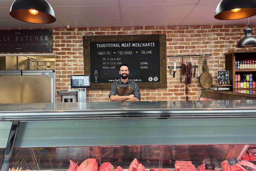 A smiling butcher with a rich dark beard stands behind the counter with his arms folded.