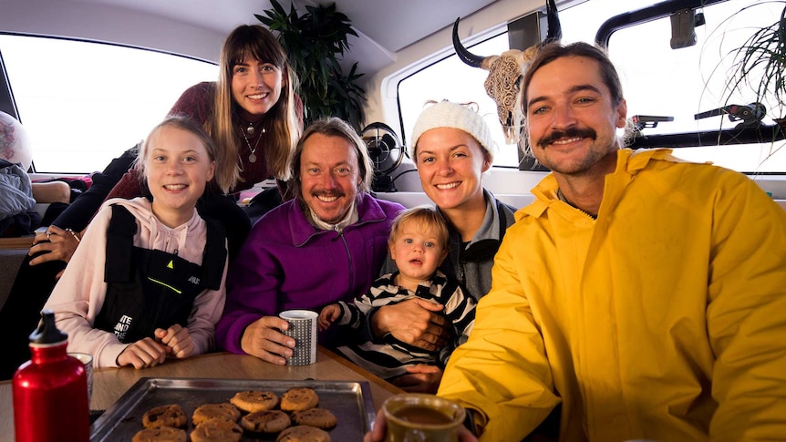 A group of people including Greta Thunberg pose for a photo smiling while drinking from coffee mugs with a tray of biscuits.