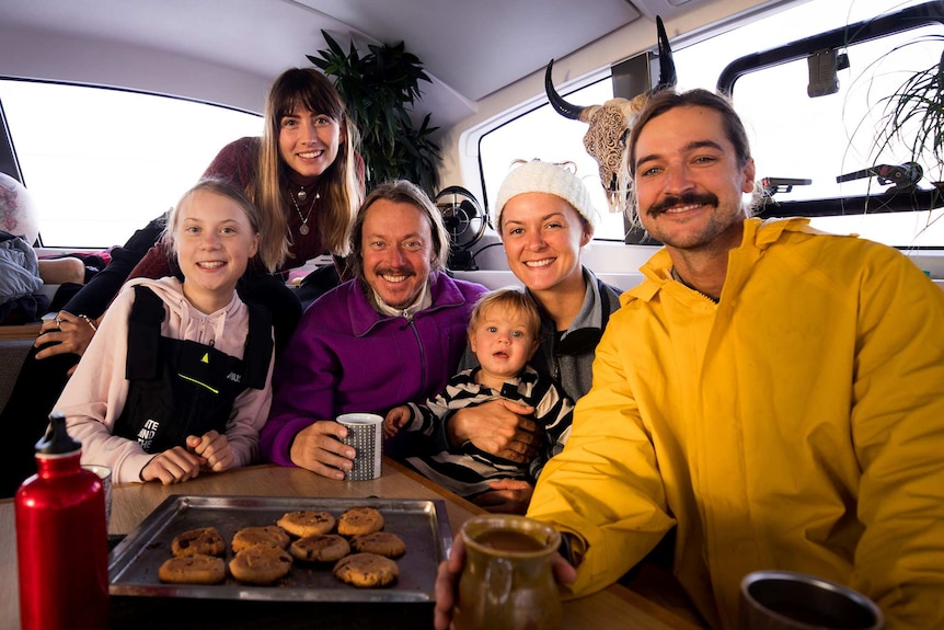 A group of people including Greta Thunberg pose for a photo smiling while drinking from coffee mugs with a tray of biscuits.