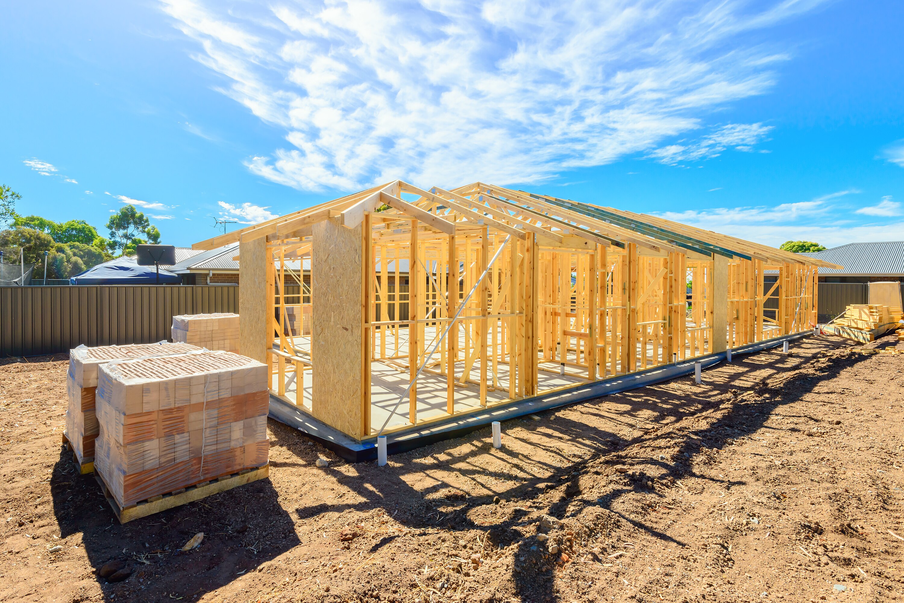 A partially constructed timber house frame sits on a block of land. There are blue skies with light clouds in the background.