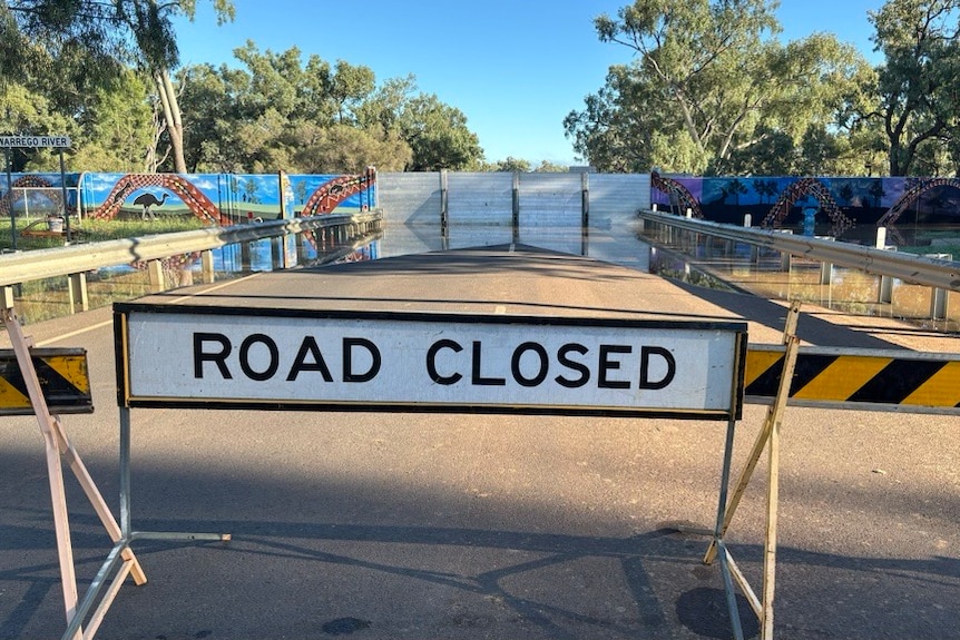 A road blocked by a ROAD CLOSED sign. Further away, water leaks under a wall to make a big puddle. 