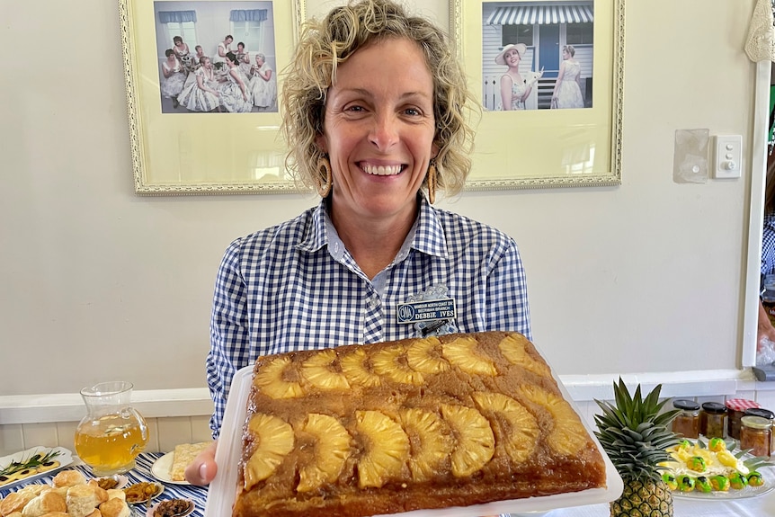 A woman holds up a big rectangular cake with slices of cooked fresh pineapple on the top.