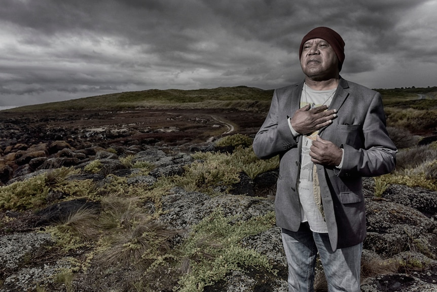 Archie Roach with black beanie, grey suit and his hand across his heart, stands smiling against a bush backdrop.
