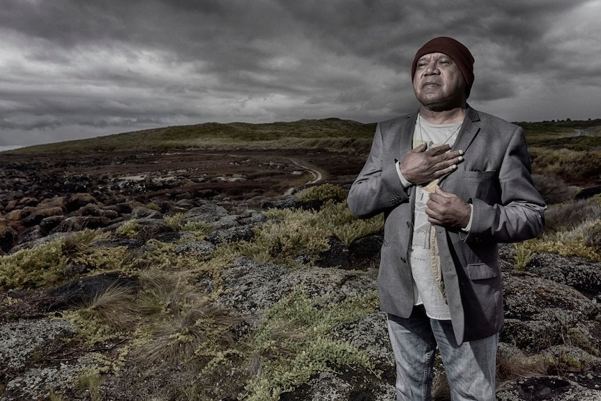 Archie Roach avec un bonnet noir, un costume gris et sa main sur son cœur, se tient souriant sur fond de brousse.