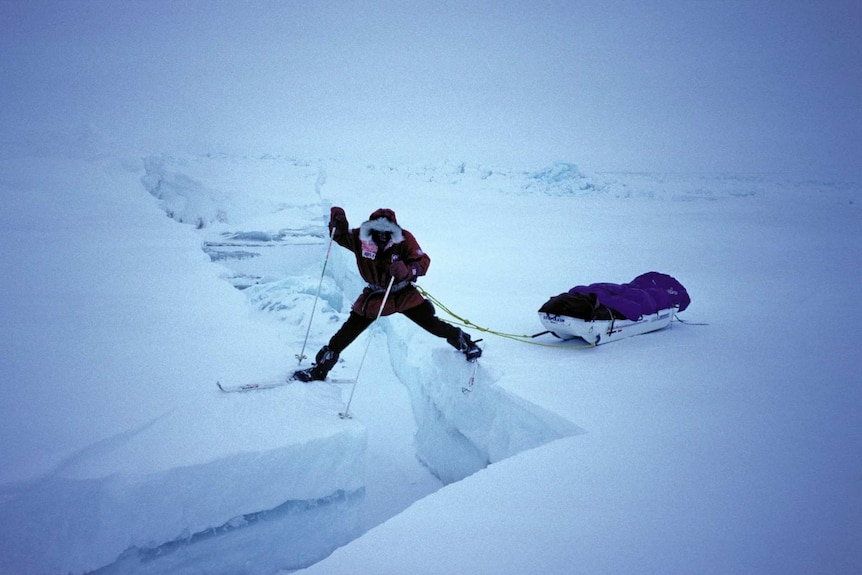 A man in a snowsuit stands with each foot on either side of a large crack in the ice.