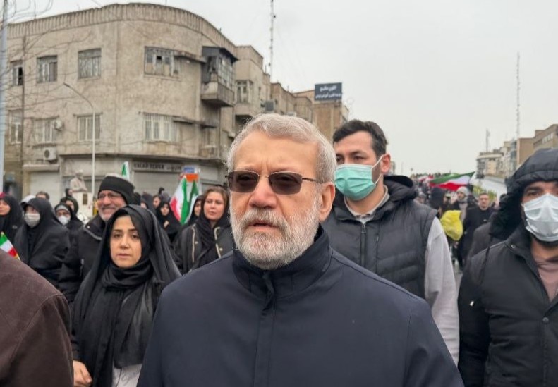 A man with a grey beard and hair walks amongst a crowd in a street. He is wearing sunglasses.
