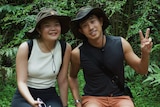 a young man and woman sit on a log wearing hats in a green forest.
