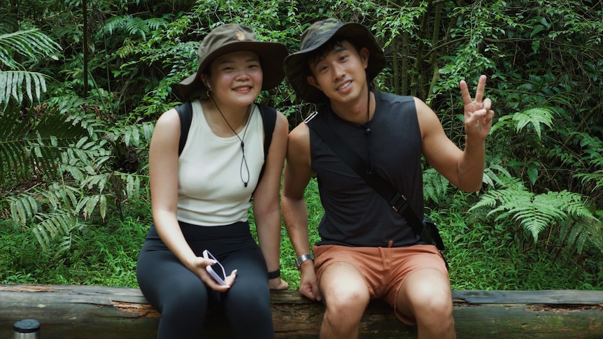a young man and woman sit on a log wearing hats in a green forest.