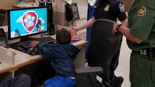 A small child is seen from behind watching Paw Patrol on a computer, while US Customs and Border Protection officers look on.