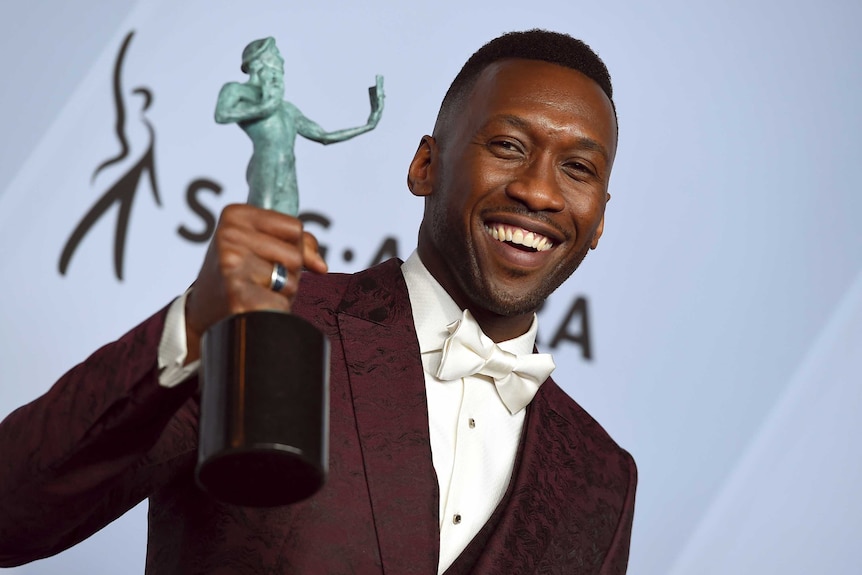 Mahershala Ali wears a maroon blazer, white shirt and black tie. He grins as he holds mint green SAG award aloft.