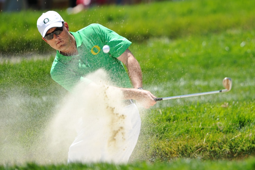 A spray of sand flies into the air as a golfer chips his ball out of a bunker.